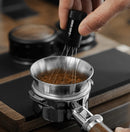 Person using a whisk to mix coffee grounds in a metal filter on a stove.