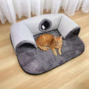An orange tabby cat lies on a plush gray pet bed with built-in arch tunnels and a circular window, placed on a light wood floor in front of white curtains.