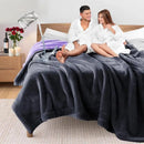 Couple in robes sitting on a bed with a dark gray blanket, surrounded by bath products.