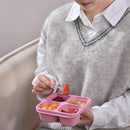 Person holding a pink compartmentalized snack tray with food, wearing a gray sweater and white shirt.